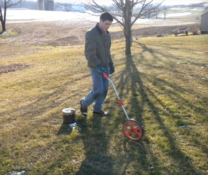 A man in a jacket and gloves using a measuring wheel on a grassy field near a covered pipe.
