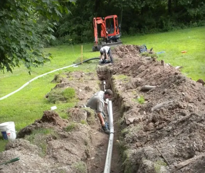 Two workers installing a long gray pipe in a narrow trench with an orange excavator in the background on a grassy area.