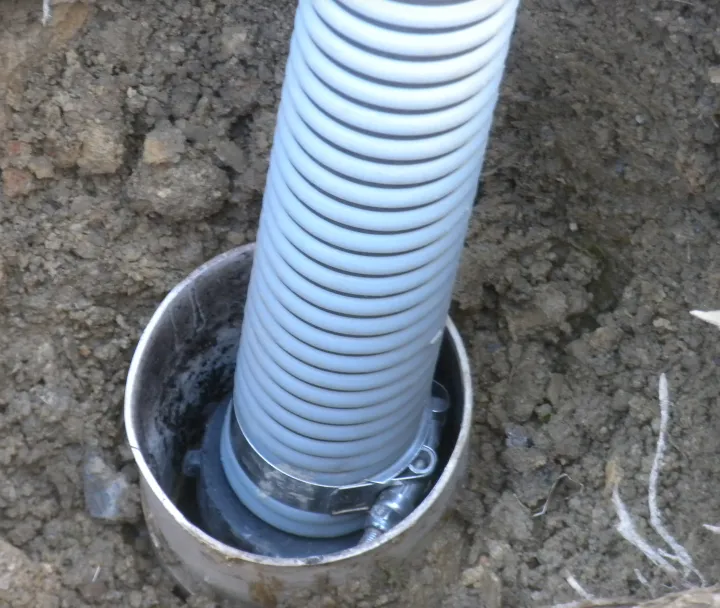 Close-up of a flexible gray corrugated pipe secured with a metal clamp inside a larger metal pipe buried in the soil.