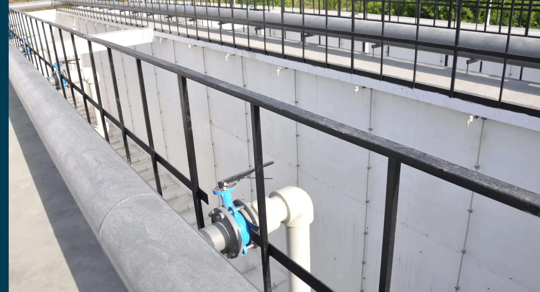 Industrial walkway with large gray pipeline and black metal railing alongside a white concrete structure.