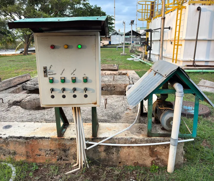 Industrial control panel with indicator lights and labeled switches next to a covered pump and piping outdoors.
