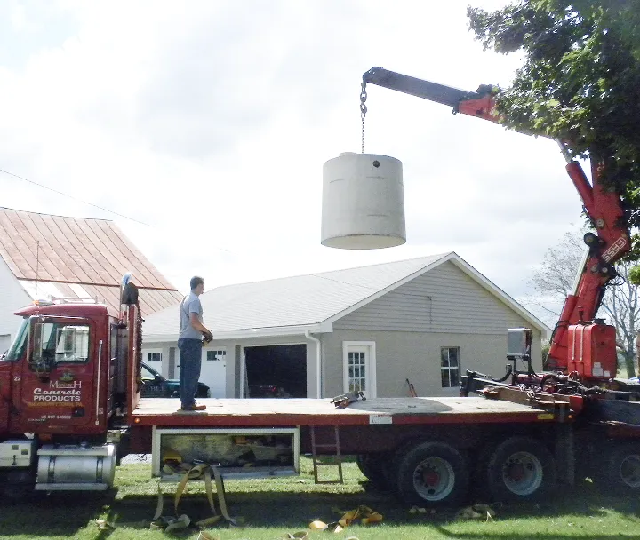 Man standing on flatbed truck watching cylindrical concrete structure being lifted by crane arm.