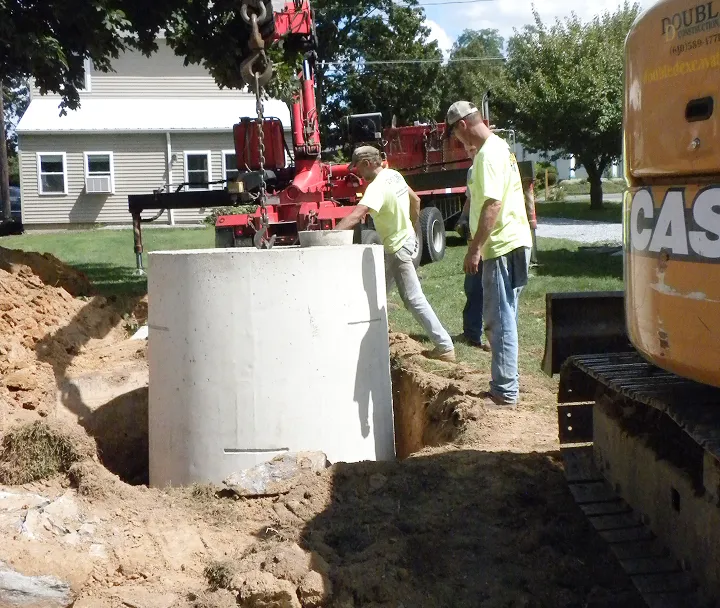 Two construction workers guiding a large concrete cylinder being lowered into a deep dug hole in a residential yard.