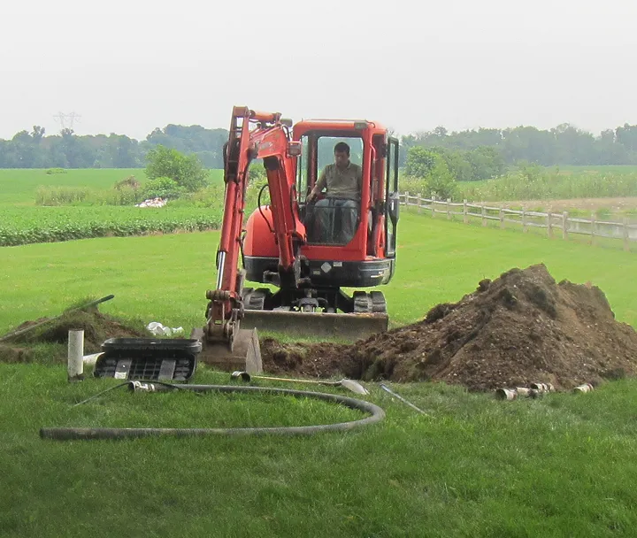 Man operating a small red excavator digging in a grassy field with a large pile of dirt and scattered pipes nearby.