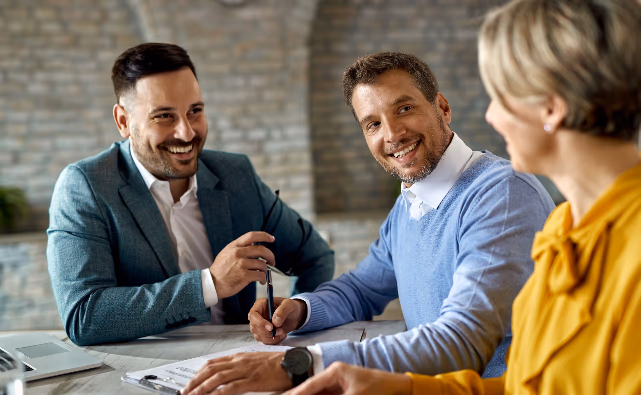 Three professionals smiling and engaging in a business meeting at a desk.
