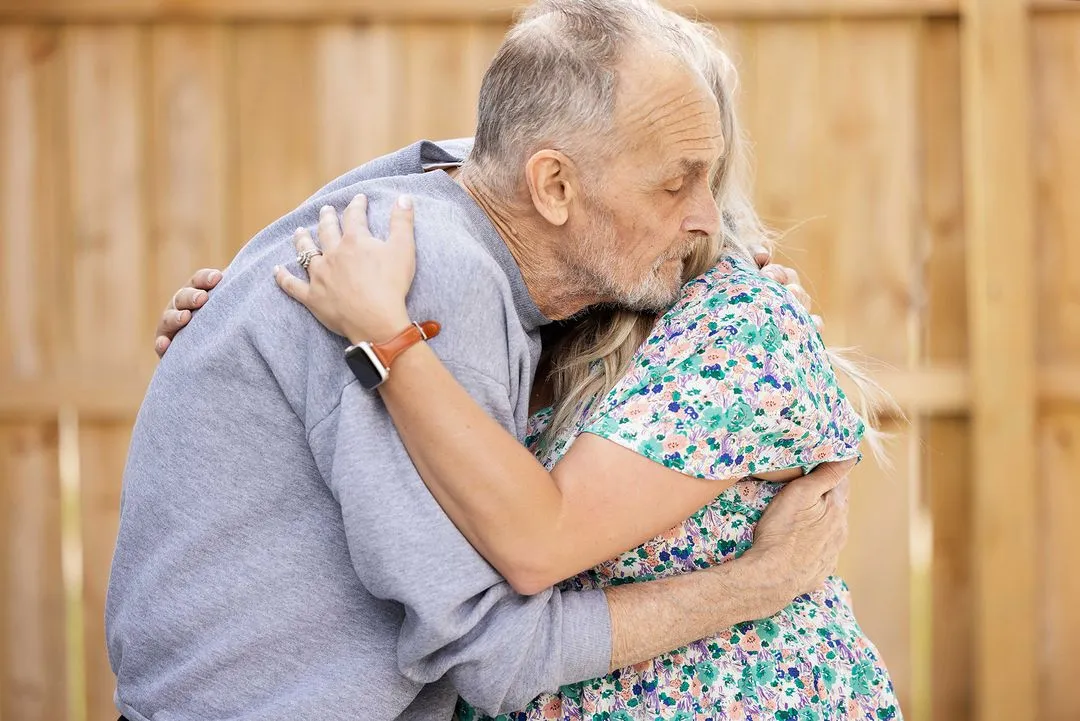 Loving staff member hugging elderly man outside in enclosed courtyard.