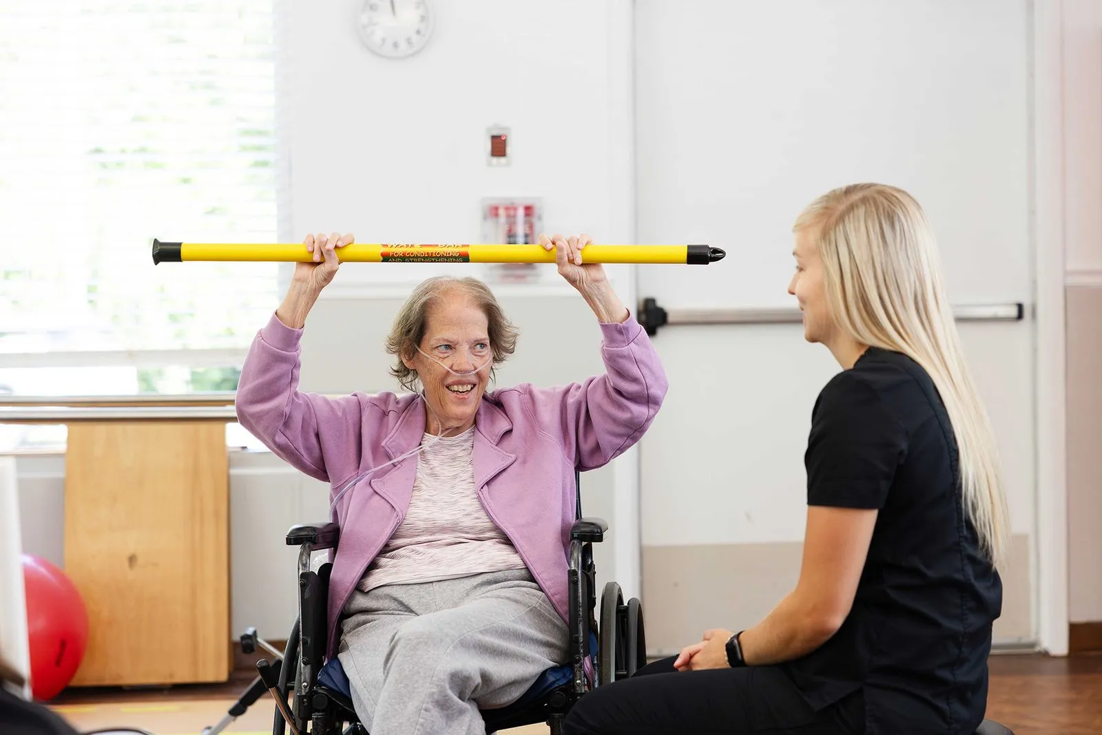 A happy elderly woman in a wheelchair does an exercise while a younger woman assists