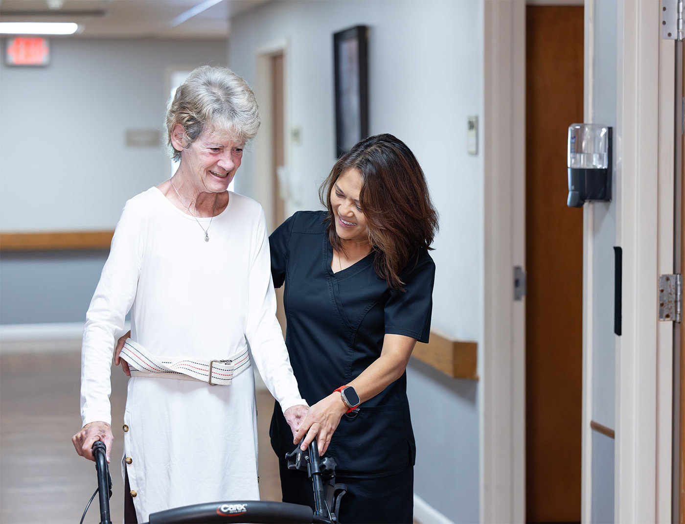 A caregiver assists an elderly woman using a walker in a hallway.