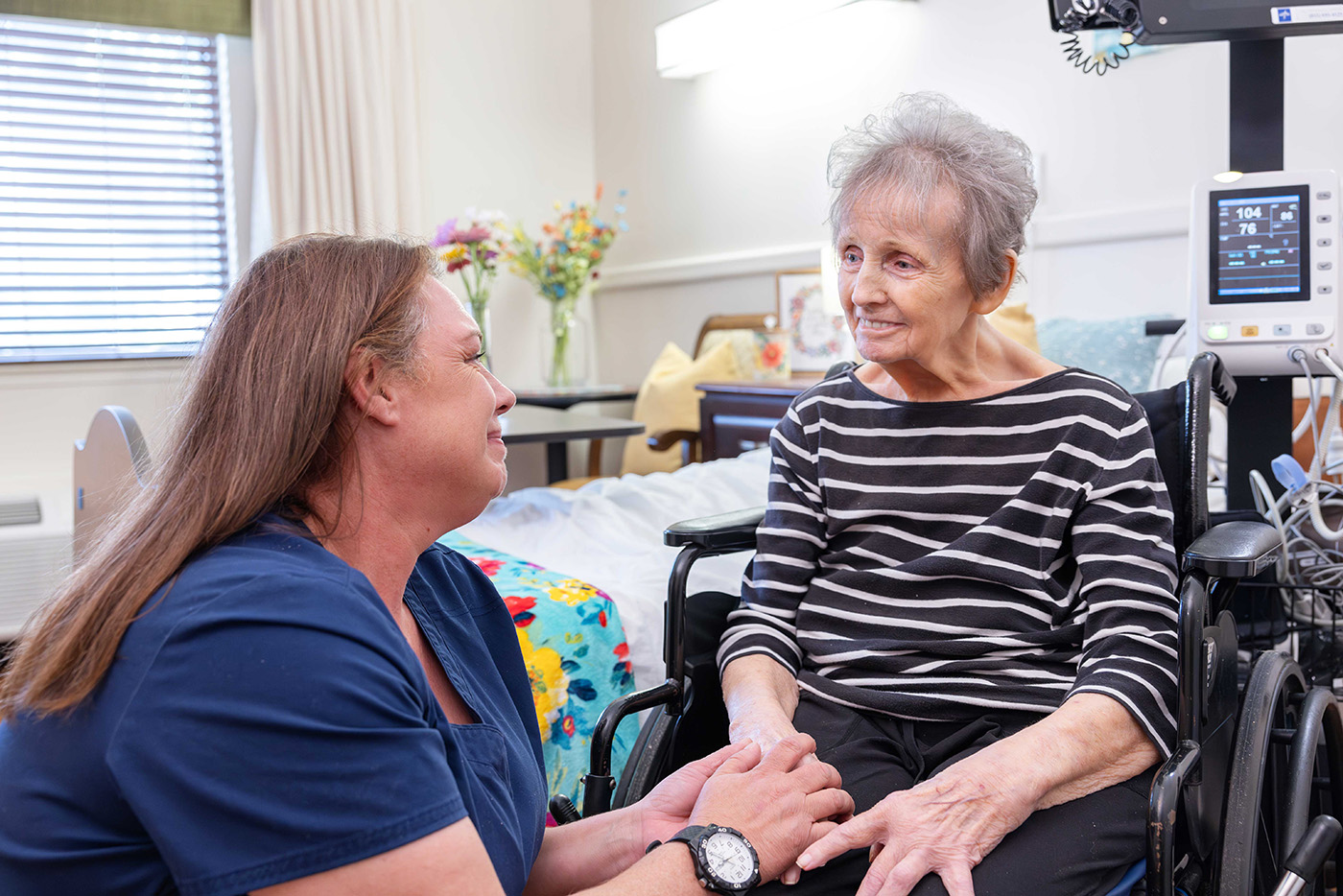 A caregiver smiles and holds hands with a woman sitting in a wheelchair.