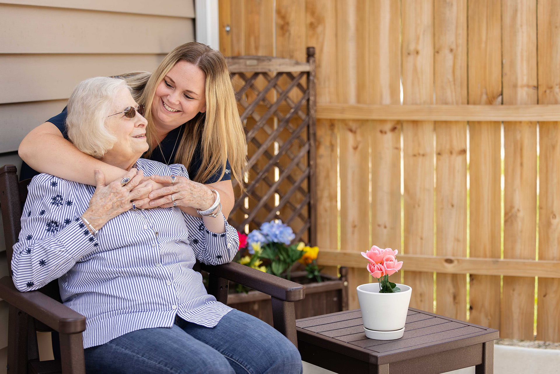 A woman hugs an older woman seated on a patio chair, both smiling warmly.
