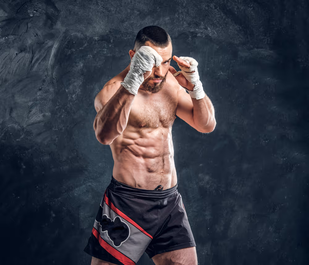 Muscular male boxer in black and red shorts with wrapped hands in a fighting stance against a dark textured background.