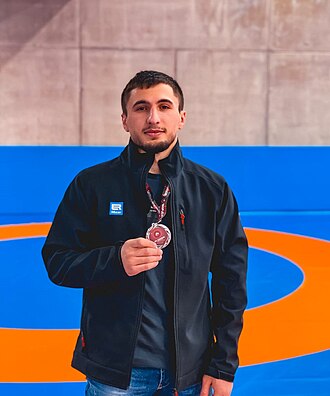 Portrait of a young man with short curly hair and a beard wearing a gray checkered shirt outdoors with green foliage background.