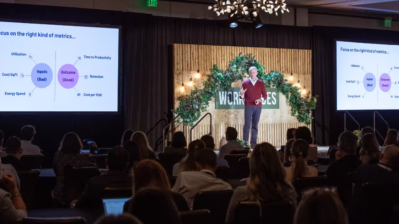 Speaker presenting on stage in front of an audience with two screens showing a diagram titled 'Focus on the right kind of metrics' comparing 'Inputs (Bad)' and 'Outcome (Good)'.
