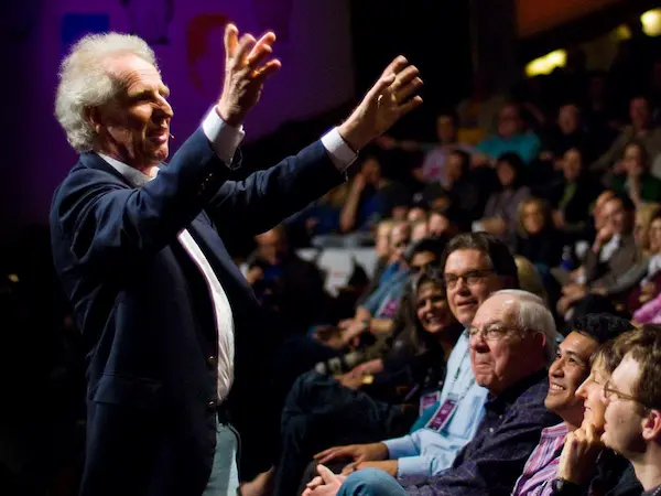 Conductor with white hair and blue suit gestures expressively to an audience seated in front of him during a presentation.