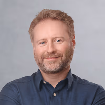 Smiling middle-aged man with light brown hair and beard wearing a dark blue button-up shirt against a gray background.