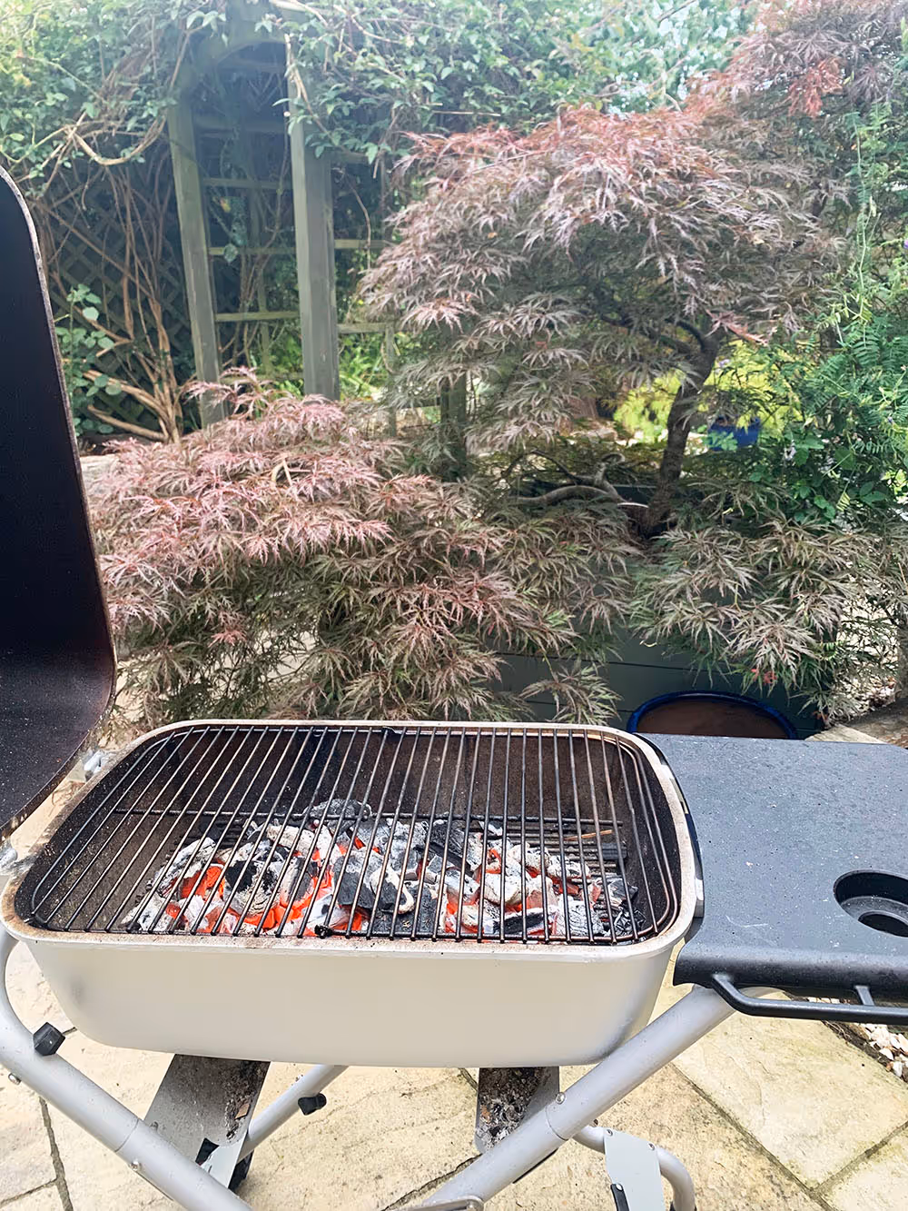 Charcoal grill with hot coals glowing, set outdoors in a garden with leafy trees in the background.