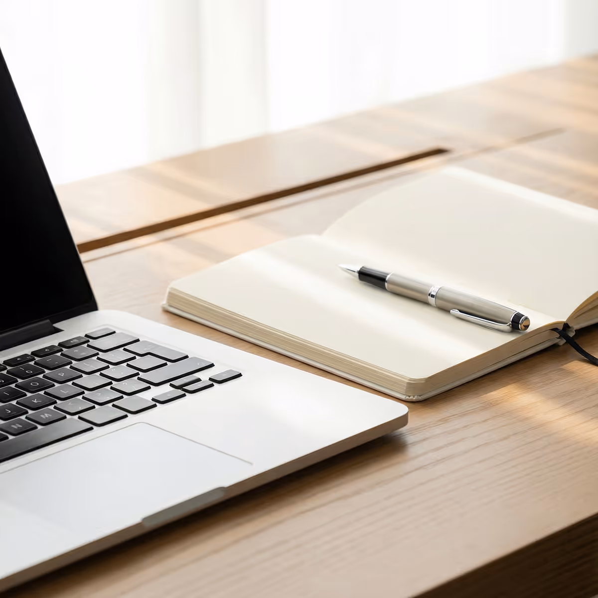 Open laptop next to an open blank notebook with a pen on a wooden desk.