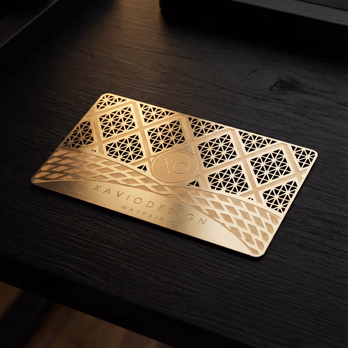 Elegant gold metal card with intricate geometric cutout patterns and 'XavioDesign Mayfair' engraved on a dark wooden surface.