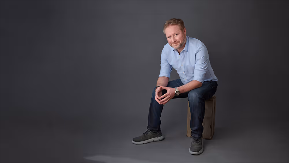 Smiling man with short hair and beard wearing a light blue shirt and dark jeans sitting on a wooden box against a gray background.