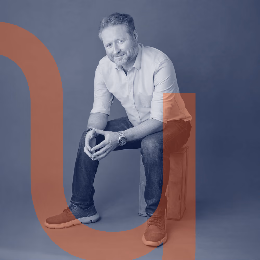 Man with light brown hair and beard wearing a light blue shirt and jeans sitting on a wooden box against a gray background.