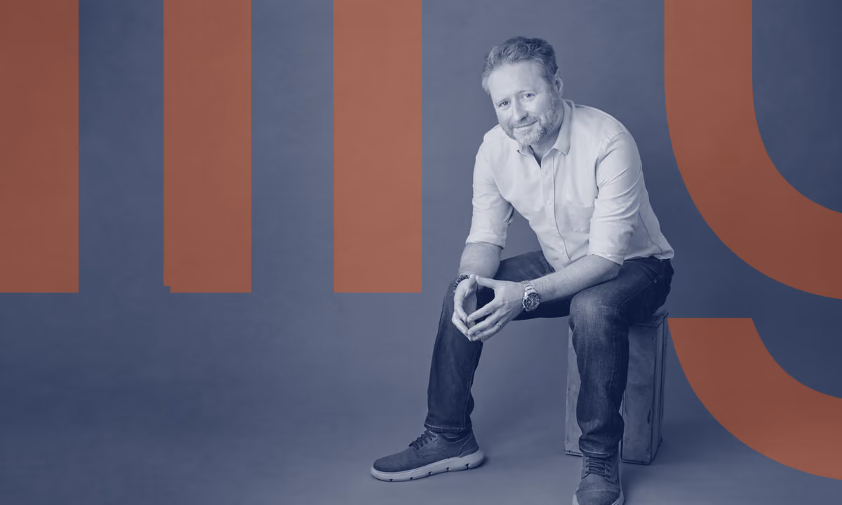 Man with light brown hair and beard sitting on a wooden box against a dark gray background, wearing a light blue shirt and dark jeans.