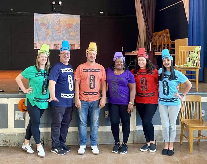 Six staff members wearing colorful crayon costumes in school auditorium