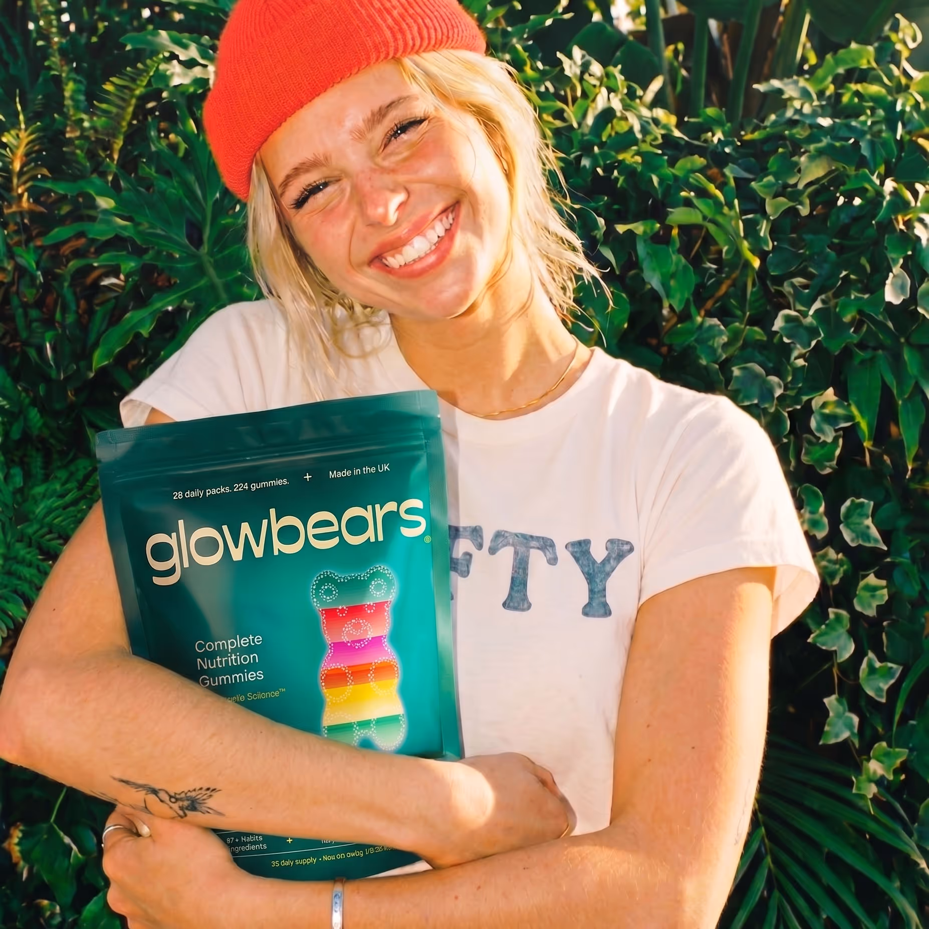 Smiling woman wearing a red beanie hugs a green pack of Glowbears Complete Nutrition Gummies in front of leafy plants.
