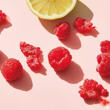 Scattered fresh raspberries and a lemon slice on a pale pink surface casting shadows.