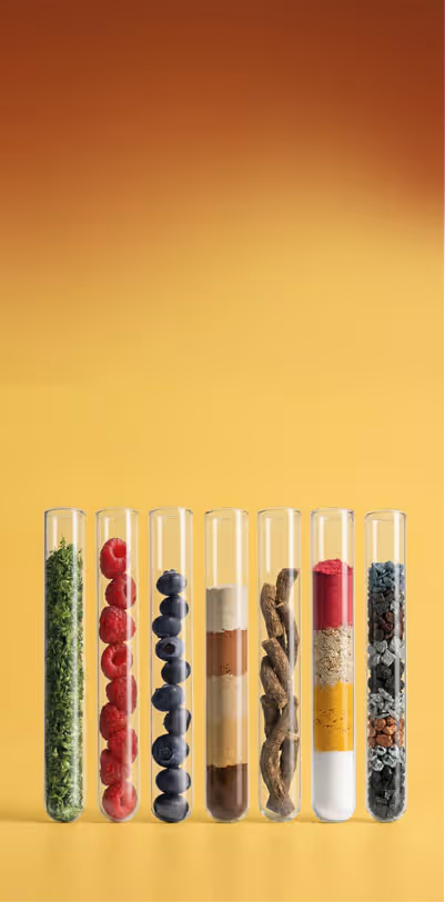 Seven clear test tubes standing upright, each filled with different colorful ingredients including green herbs, red berries, blueberries, layered powders, twisted roots, and various granules, against a yellow background.