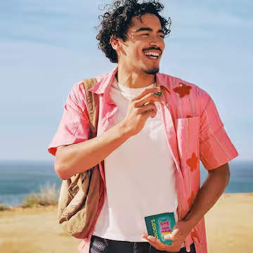 Smiling man with curly hair wearing a pink shirt and backpack, holding a pack of gummy vitamins outdoors near the ocean.