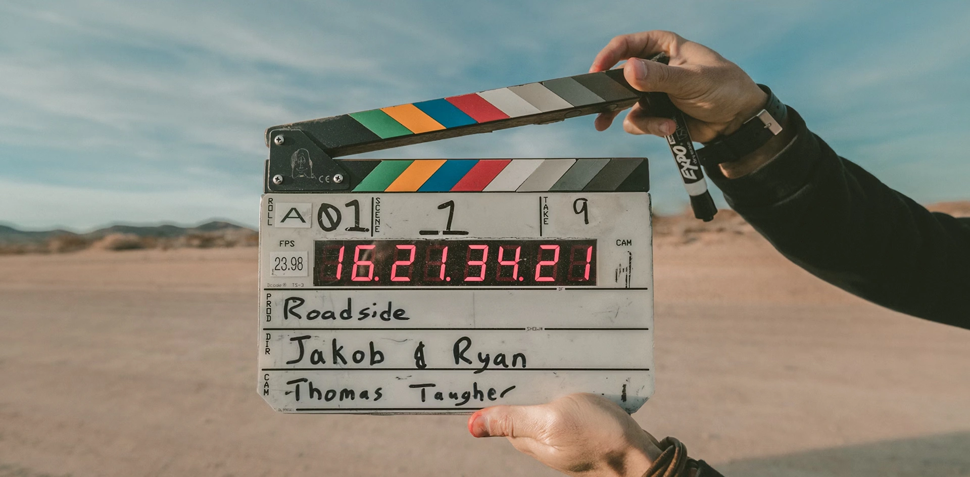 Clapperboard held by hands in a desert setting showing production details and timecode.