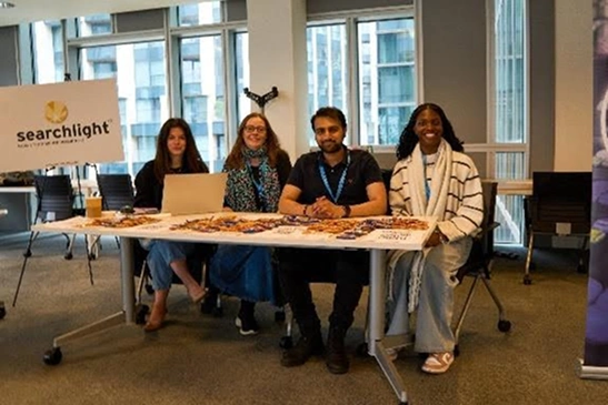 Four people sitting behind a table with promotional materials and a Searchlight sign in a modern office setting.