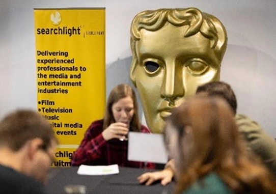 Large golden BAFTA mask beside a yellow Searchlight recruitment banner and people sitting at a table in discussion.