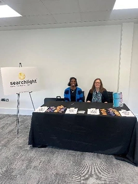 Two women sitting behind a table covered with promotional items and flyers, next to a sign that reads 'Searchlight'.