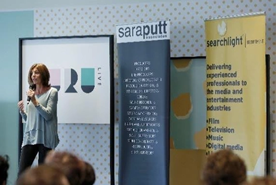 Woman speaking with microphone in front of banners for Saraputt Associates and Searchlight, addressing an audience.