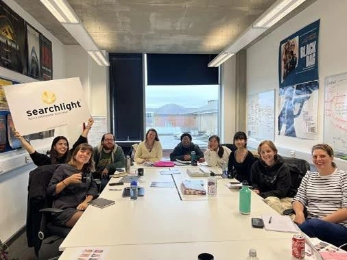Group of nine people sitting and smiling around a conference table in a meeting room, one person holding a 'searchlight' sign.