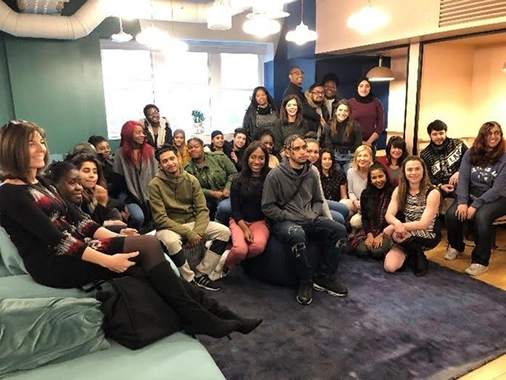 Group of diverse young adults sitting and standing together in a cozy, modern room with blue walls and pendant lights.