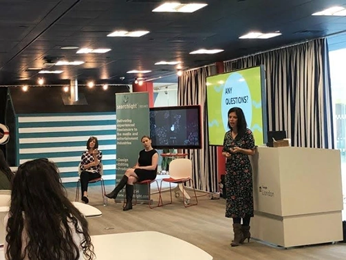 Woman standing and speaking next to a podium in a conference room while two seated women and an audience member listen.
