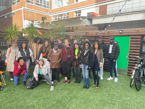Group of diverse young adults posing outdoors on artificial grass in front of a building and wooden fence.
