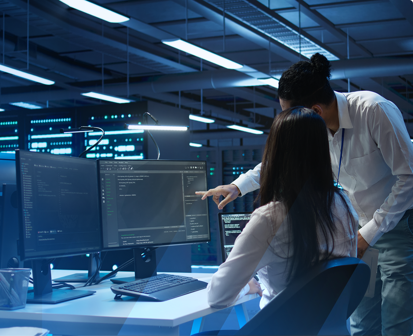 Two IT professionals collaborating in a data center, with multiple monitors displaying code and server racks in the background.