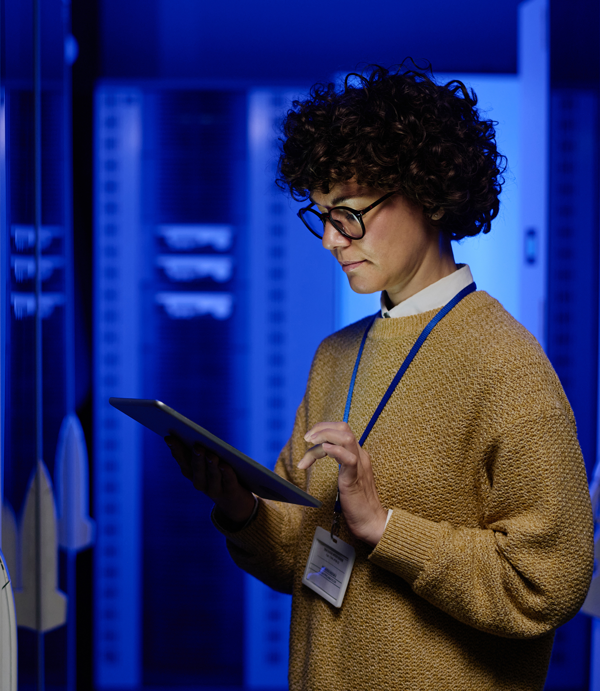 Woman with curly hair and glasses working on a digital tablet in a blue-lit server room.