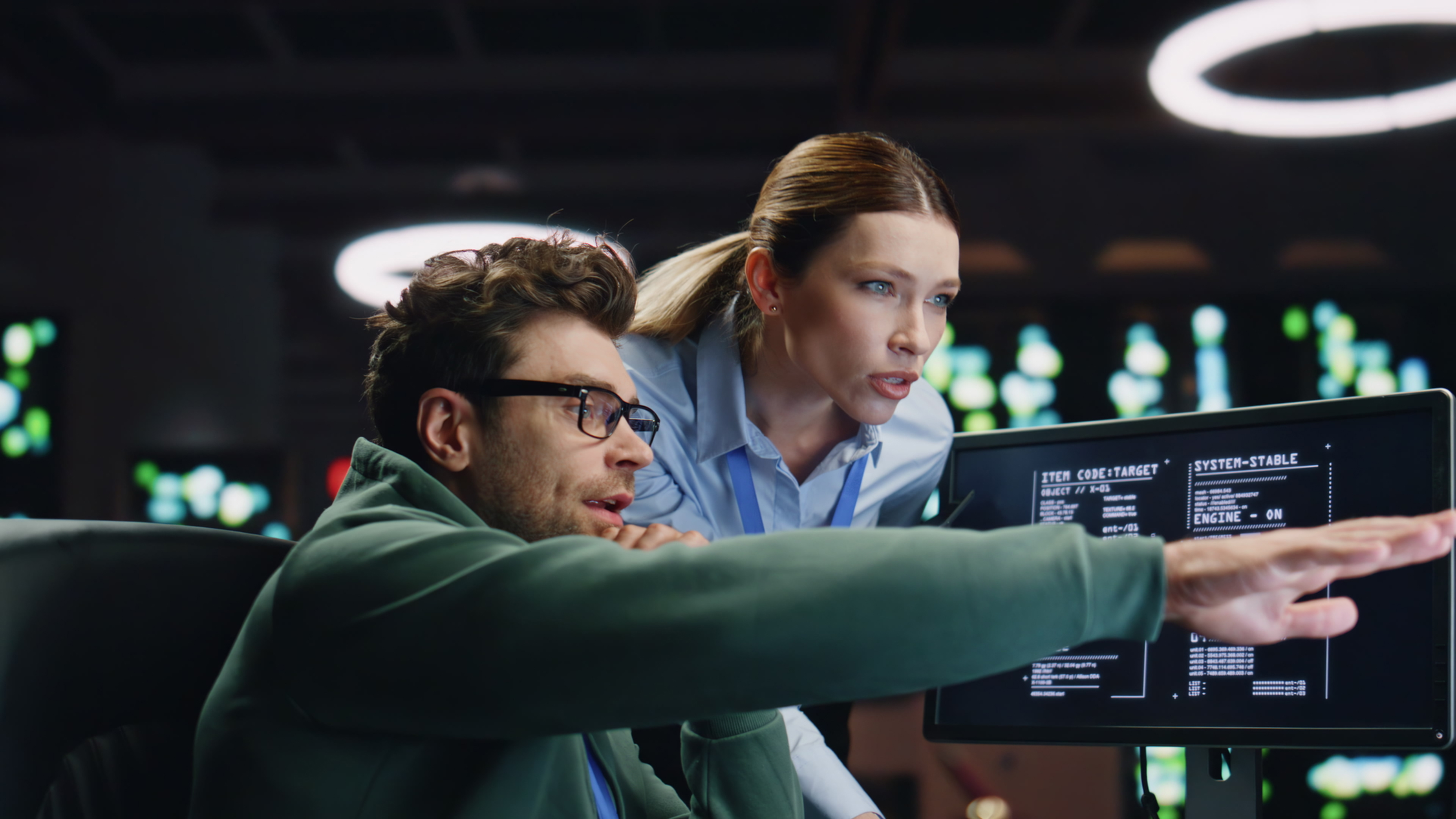 Two programmers focused on a computer screen displaying code in a dark room, with one pointing at the monitor.