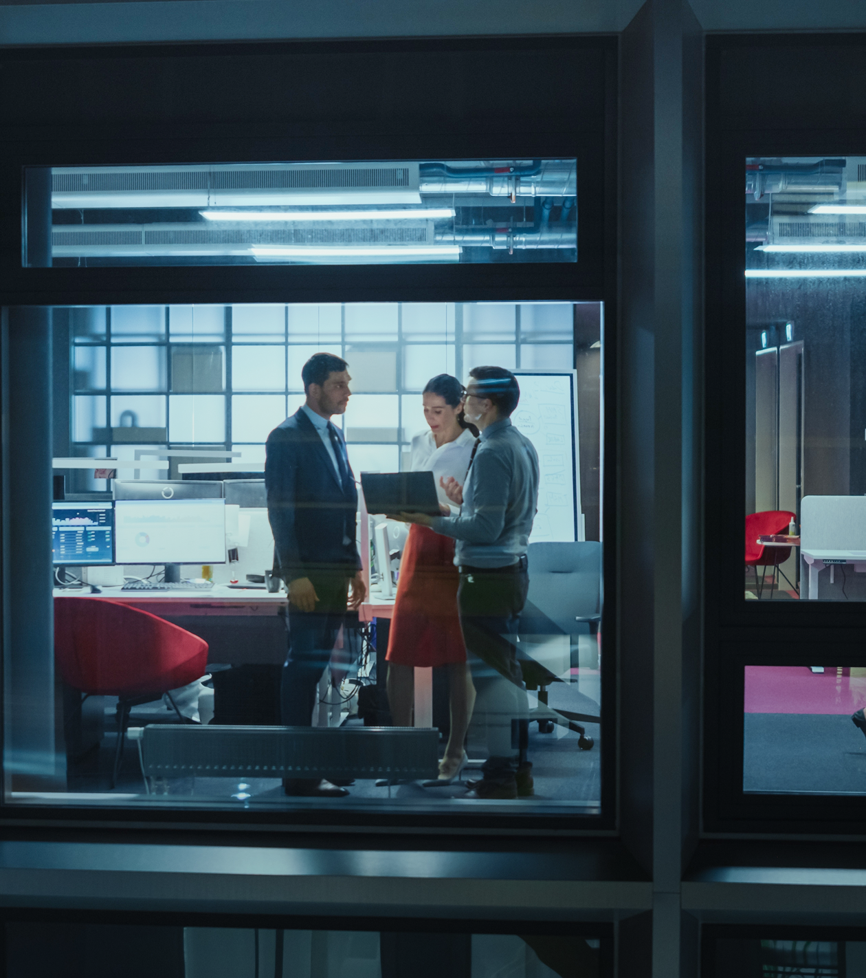 Three business professionals standing and discussing a laptop in a modern office with computer monitors and a whiteboard.