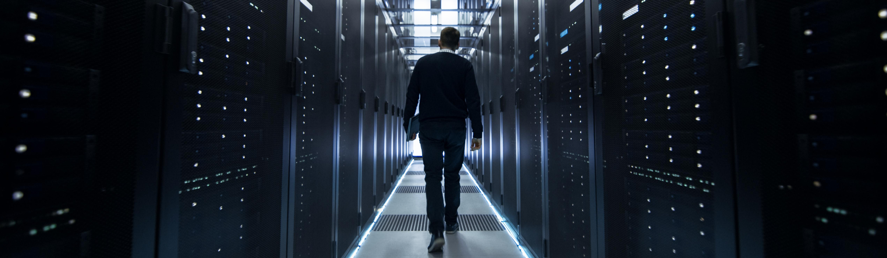 Man walking down aisle between rows of illuminated server racks in a data center.