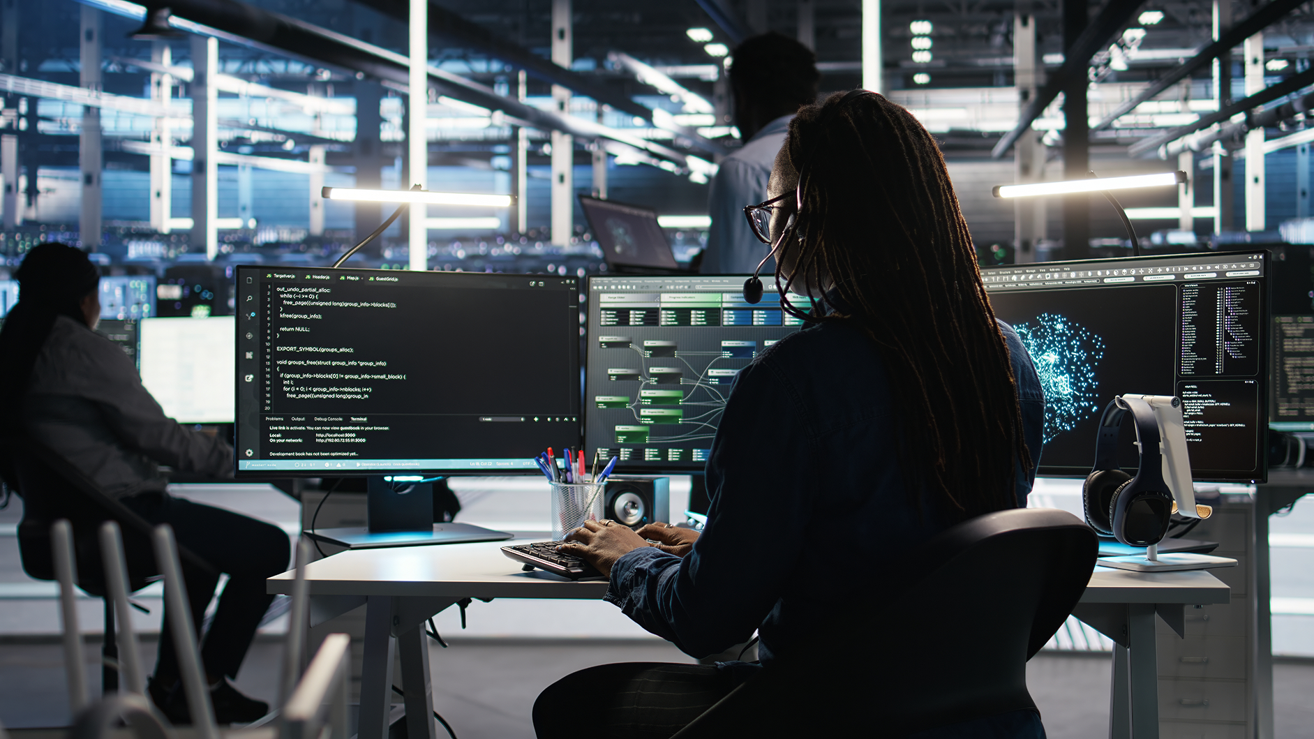 Person with dreadlocks wearing headset and glasses working on multiple monitors with code and data visualizations in a modern office.