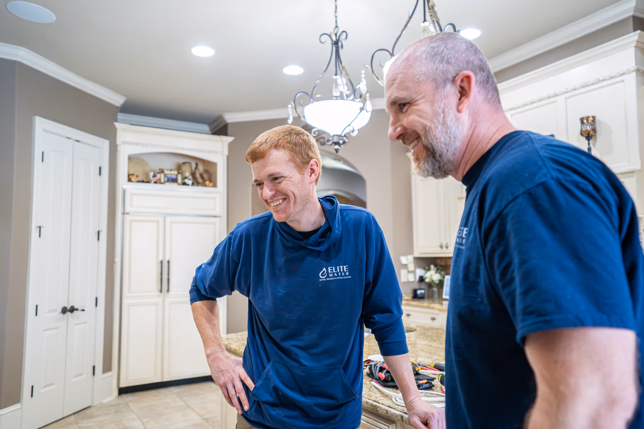 Two men in blue Elite Water shirts examining a water filtration device on a kitchen counter.