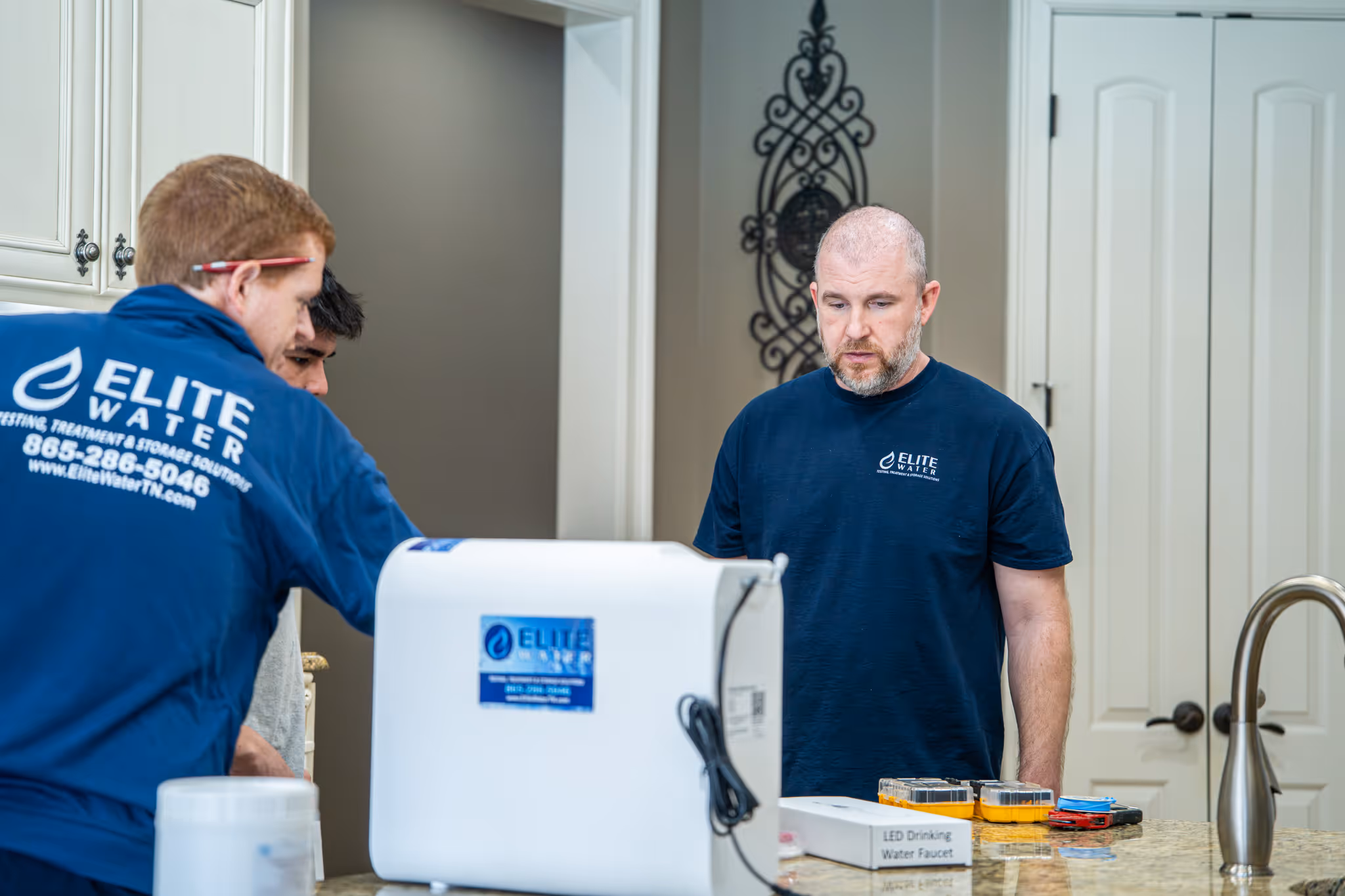 Man in a blue Elite Water hoodie crouching on grass, handling water testing equipment outdoors near trees and a parked white truck.