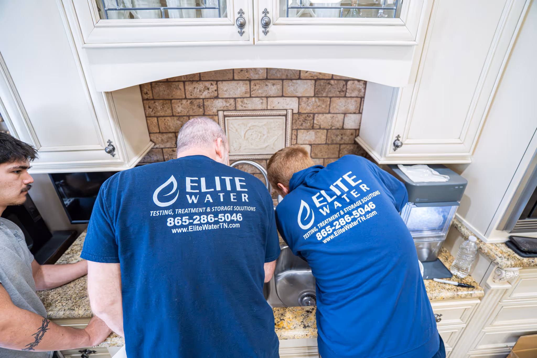 Two Elite Water technicians in branded shirts working together at a kitchen sink while a man watches.