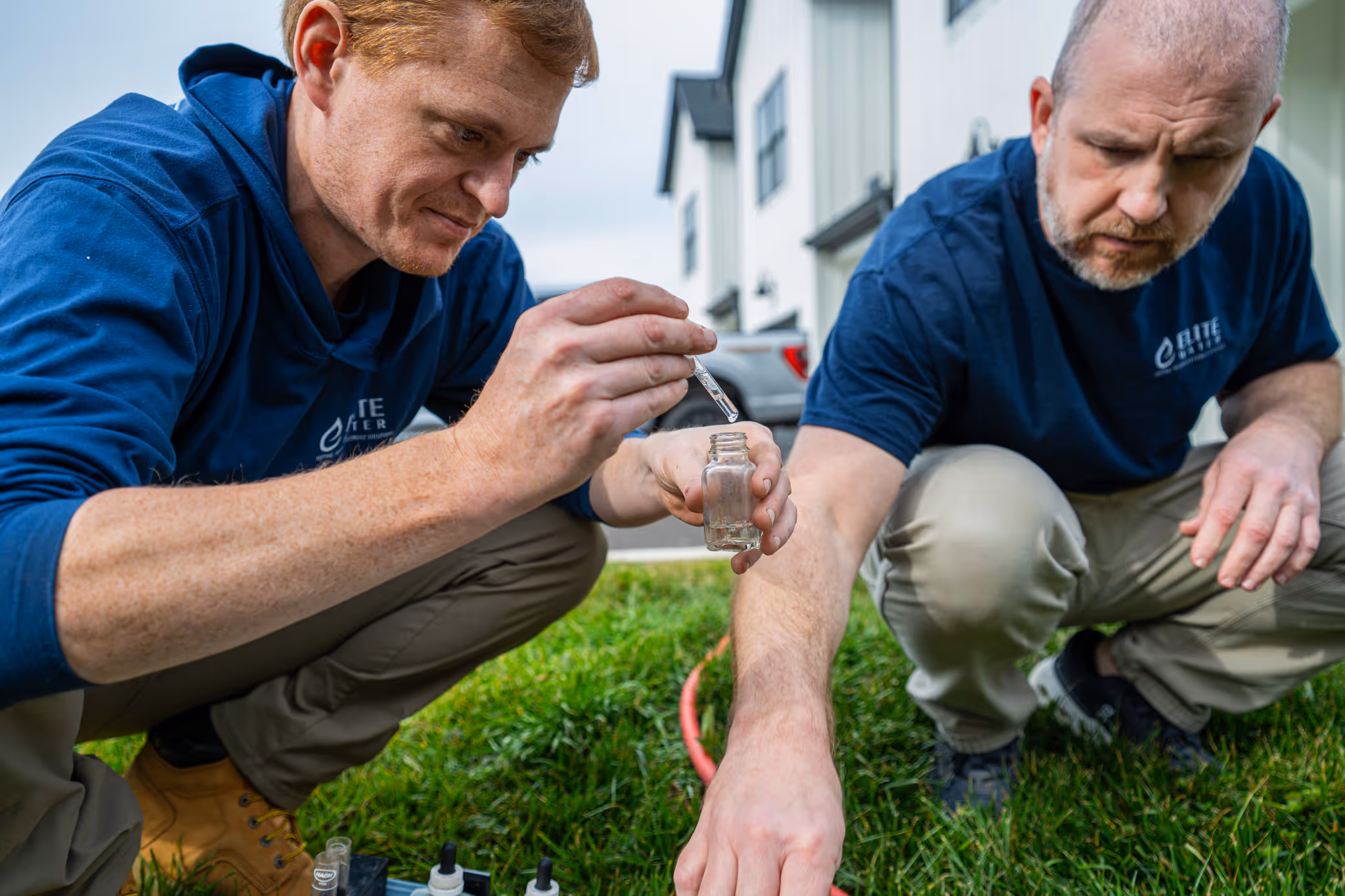 Two men in blue shirts crouching on grass outdoors, one holding a dropper and a small glass container for water testing.
