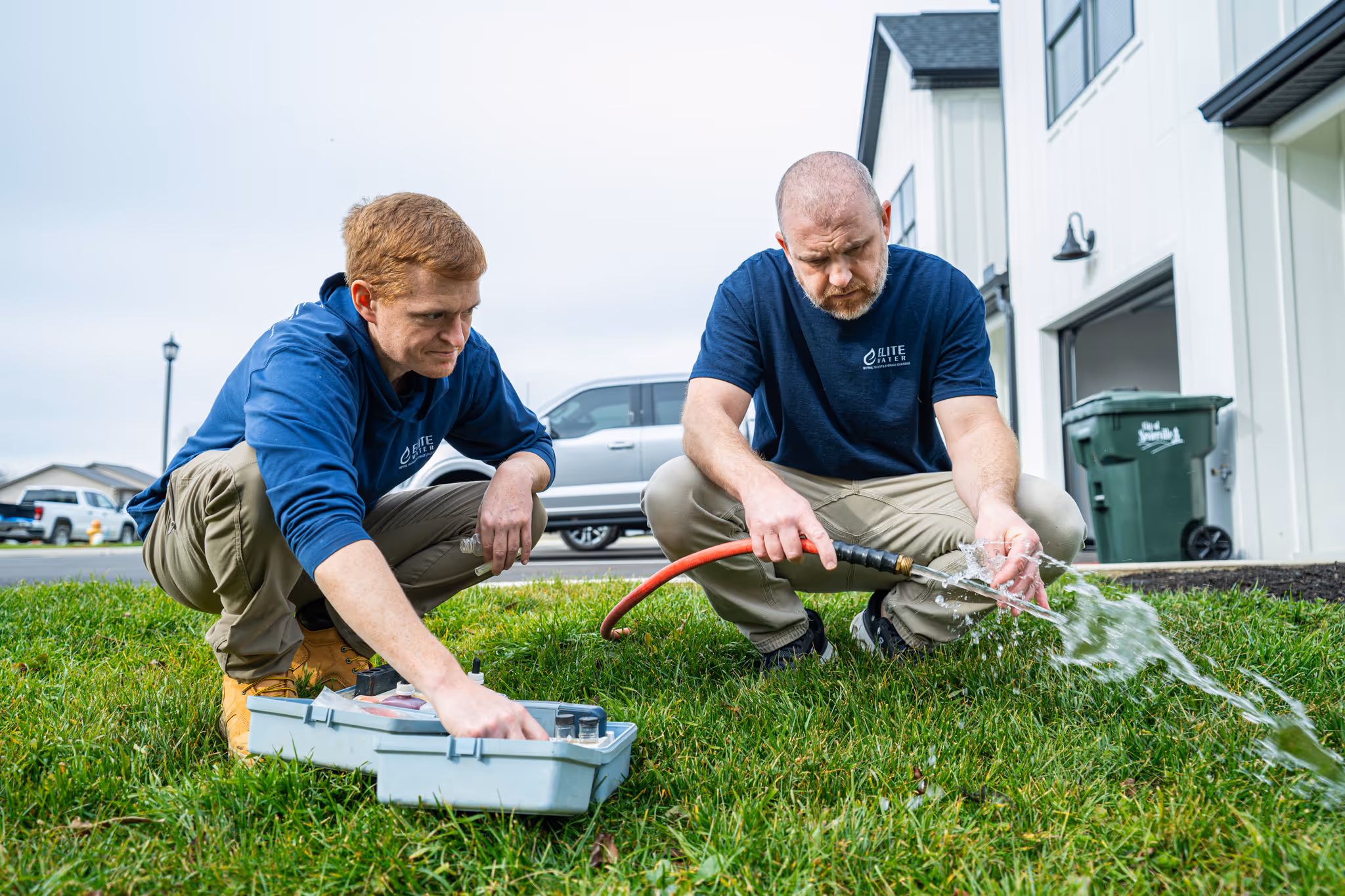 Two men in blue Elite Water shirts working on a water hose and tools on grass outside a house.